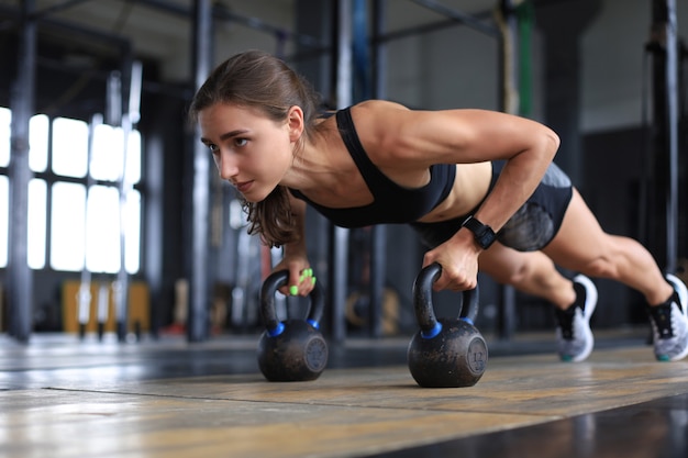 Portrait of a muscular woman on a plank position with kettlebell at gym.