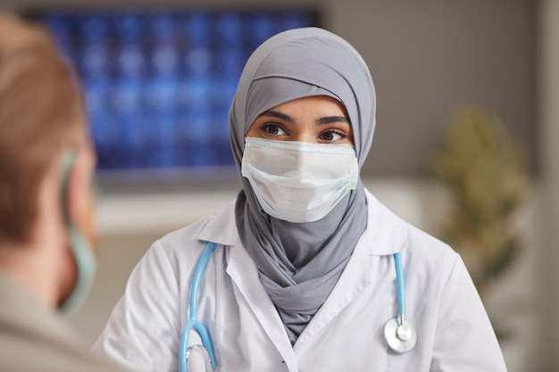 Photo portrait of muslim doctor in protective mask looking at her patient while they sitting at hospital
