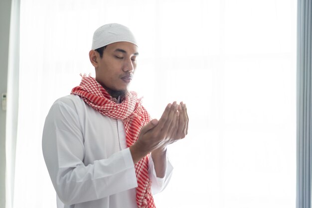 Portrait of muslim man using hand to pray against white window