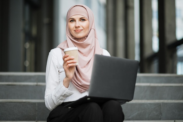 Portrait of muslim woman in hijab working on laptop outdoors