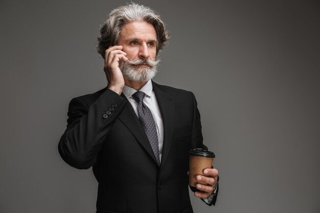 Portrait of mustached adult businessman wearing formal black suit holding paper cup and talking on smartphone isolated over gray wall