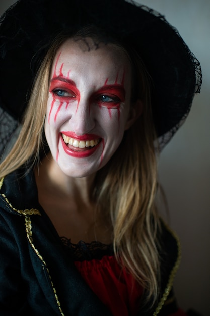 Portrait of a mysterious girl with fair hair as a witch with bright make-up and leaking lipstick mysteriously looks to the side with wide smile.