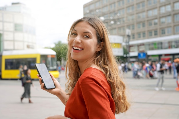 Портрет натуральной красивой улыбающейся женщины, идущей по берлинской площади Александерплац с мобильным телефоном на размытом городском фоне