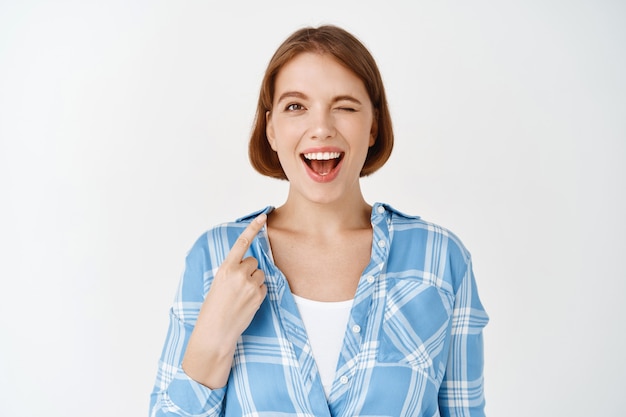 Portrait of natural happy girl winking, smiling and pointing at herself, self-promoting, talking about her achievement, showing skills, standing on white wall