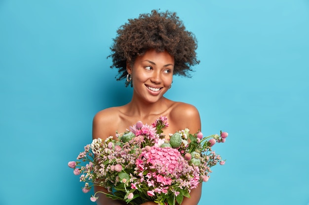 Portrait of natural young woman looks aside gladfully smiles gently holds beautiful boquet over naked body poses with bare shoulders isolated