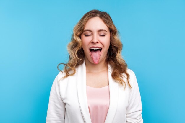 Portrait of naughty positive woman with wavy hair in white jacket standing with closed eyes and demonstrating tongue, expressing disobedience disrespect, teasing grimace. studio shot, blue background
