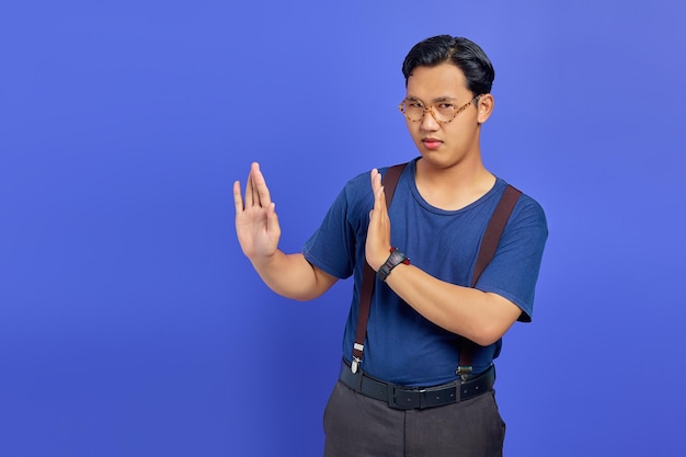 Portrait of nervous anxious young asian man make stop don\'t\
move gesture over purple uebackground