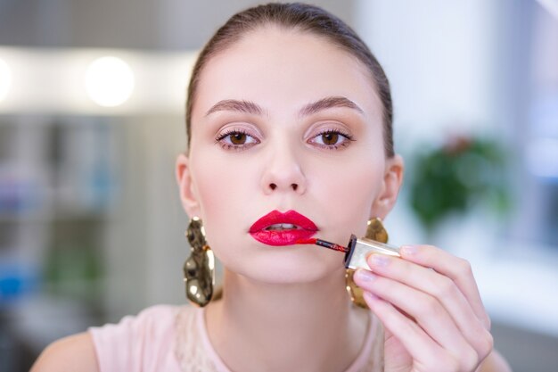 Portrait of a nice attractive woman looking at you while putting on lipstick