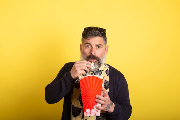 Portrait of nice attractive worried scared addict bearded man with yellow shirt and blue jacket eating popcorn snack watching scary video isolated on yellow background.