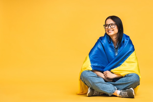 Portrait of nice beautiful lovely glad cheerful woman holding in hands Ukrainian flag having fun isolated over yellow pastel color background Sitting on the floor in lotus pose