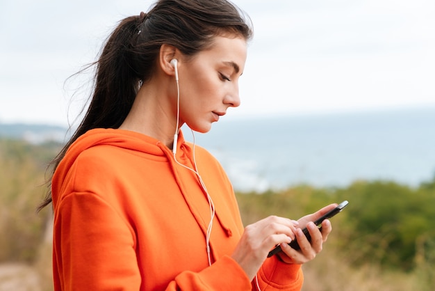 Photo portrait of nice brunette woman in sportswear using earphones and cellphone while working out at seashore