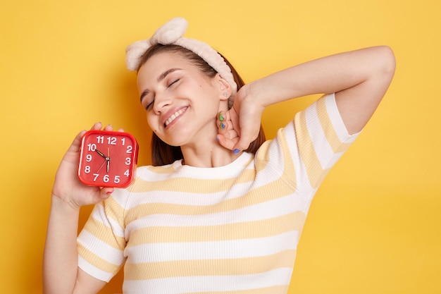 Portrait of nice cute charming winsome attractive woman wearing striped tshirt holding in hand red alarm clock statching hands wakes up in good mood posing isolated over yellow background