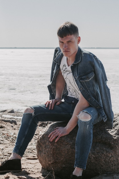 Portrait of a nice-looking serious young man sitting on a stone on a cold weather in a jeans outfit.