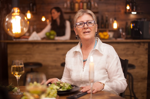 Portrait of nice old lady looking at the camera while enjoying her meal in a restaurant. A glass of white wine. Salad of vegetables.