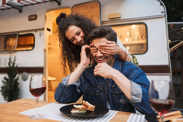 Portrait of nice smiling woman with curly long hair covering her boyfriend‘s eyes at dinner near trailer outdoors