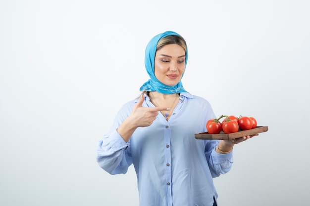 Portrait of nice woman in blue shawl pointing at wooden board of red tomatoes 