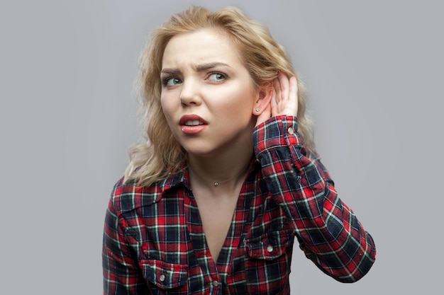 Portrait of nosy beautiful blonde young woman in casual red checkered shirt standing with hand near ear and trying to hear and spying. indoor studio shot, isolated on grey background.