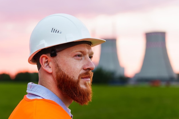 Portrait of a nuclear power plant engineer A man with a beard in a protective helmet and an orange vest stands against the background of a nuclear power plant