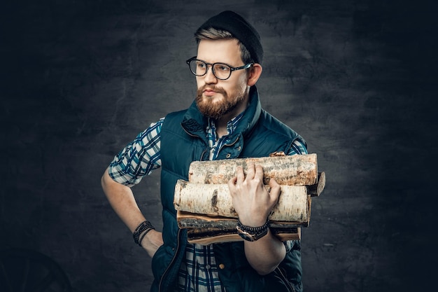Портрет бородатого мужчины, одетого в флисовую рубашку и теплый жилет, держит в руках дрова.