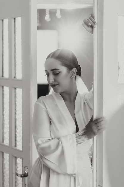 Фото Портрет красивой невесты с стильными волосами и макияжем в белом платье, позирующей, глядя в линзу возле окна балкона черно-белая фотография
