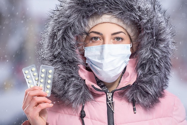 Фото Портрет красивой девушки в розовой куртке в медицинской защитной маске с желтыми таблетками в руках от простуды и гриппа зимний уличный портрет женщины под снегопадом