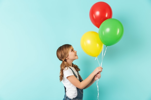 Фото Портрет веселой маленькой девочки, изолированной над синей стеной, держащей в руках кучу разноцветных воздушных шаров