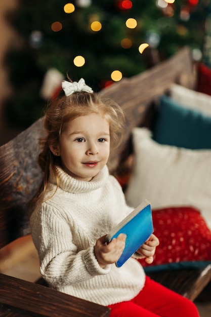 写真 クリスマス新年の家で居心地の良い冬の夜を楽しんでいるクリスマス ツリーの背景に本を手に持った暖かいニットのセーターを着た巻き毛の少女の肖像画