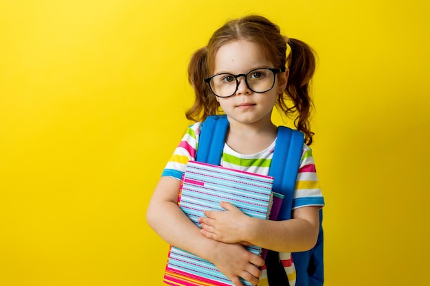 Фото Портрет милой маленькой девочки в очках в полосатой футболке с тетрадями и учебниками в руках и рюкзаком. концепция образования. фотостудия, желтый фон, место для текста.