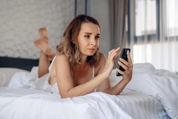 밝은 방에 있는 넓은 흰색 침대에 누워 있는 전화를 바라보는 금발의 매력적인 성적인 여성의 초상화. 아름다움의 개념