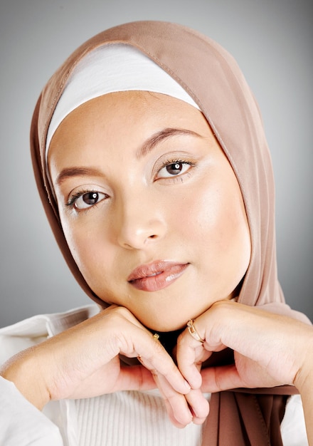 회색 스튜디오 배경에 격리된 빛나는 아름다운 이슬람 여성의 초상화 히잡이나 머리 스카프를 두른 젊은 여성이 속눈썹 연장과 장신구를 보여주는 완벽한 피부를 만지고 있다