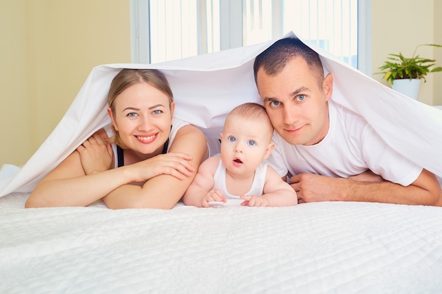 Фото Портрет счастливой семьи в комнате на кровати, мама, папа и мальчик, лежащие на кровати, спрятались с улыбкой на одеяле