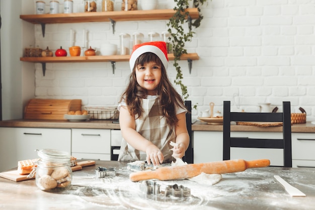 크리스마스 쿠키를 준비하는 아이가 식탁에 반죽을 굴리는 산타 모자를 쓴 행복한 어린 소녀의 초상화