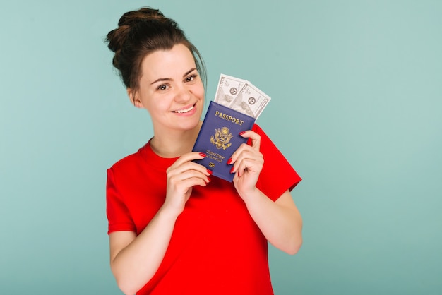 Фото Портрет счастливой улыбающейся молодой женщины, держащей паспорт и деньги в долларах сша, изолированные на синем пространстве