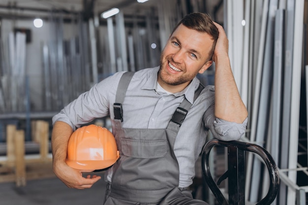 Фото Портрет счастливого рабочего в оранжевой каске и комбинезоне, держащего гидравлический грузовик на фоне завода и алюминиевых рам