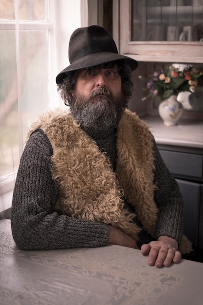 Фото Портрет мужчины, сельчанина, брюнетки в шляпе и с бородой сидит в своем доме у окна за столом