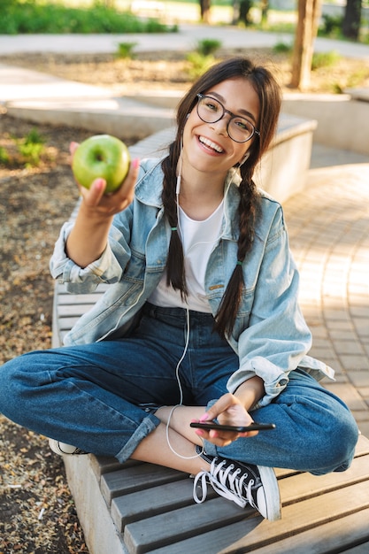 自然公園の屋外のベンチに座って眼鏡をかけている前向きで陽気な若い学生の女の子の肖像画は、リンゴを持っているイヤホンで音楽を聴いておしゃべりする携帯電話を使用しています。