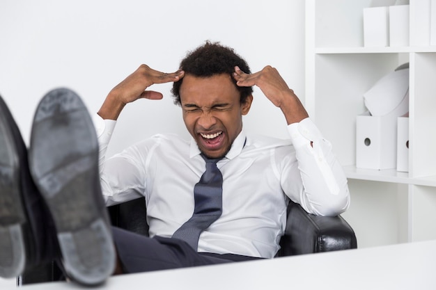 Фото Портрет усталого афроамериканского бизнесмена, у которого ужасная головная боль и стресс. он сидит в своем кабинете с ногами на столе и кричит.