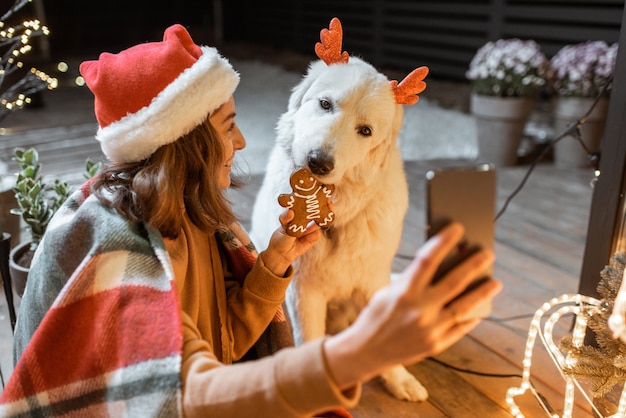 クリスマスの帽子をかぶった女性と、家で年末年始を祝うかわいい犬と格子縞の女性の肖像画、ジンジャーブレッドクッキーを犬に与え、自分撮り写真を作成する