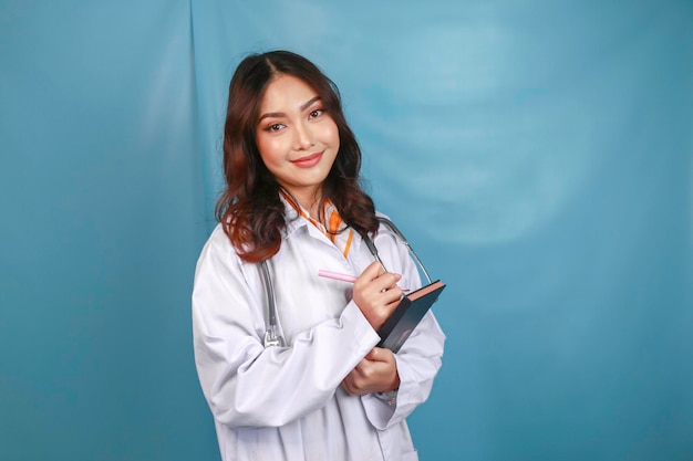 Фото Портрет молодой азиатской женщины-врача, медицинский работник улыбается и держит заметки, изолированные на синем фоне