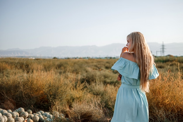 日没時に天日干しの草でスイカ畑に立っている水色のドレスで若い美しい白人ブロンドの女の子の肖像画