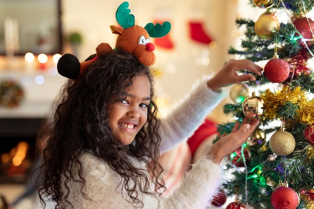 Фото Портрет молодой девушки смешанной расы в гостиной на рождество, в праздничной шляпе, держащей безделушку на рождественской елке и улыбающейся