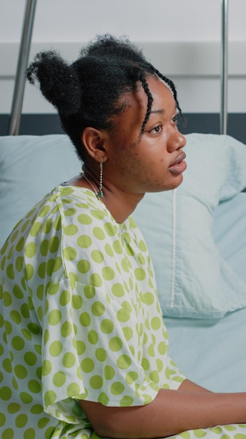病棟のベッドに座って、病気を治すための医療支援と治療を行うために医師を待っているアフリカ系アメリカ人の患者の肖像画。気分が悪いivドリップバッグを持つ若い女性