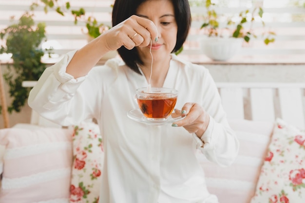 사진 그녀의 집 테라스에서 유리 컵에 차를 마시는 성인 아시아 여성의 초상화