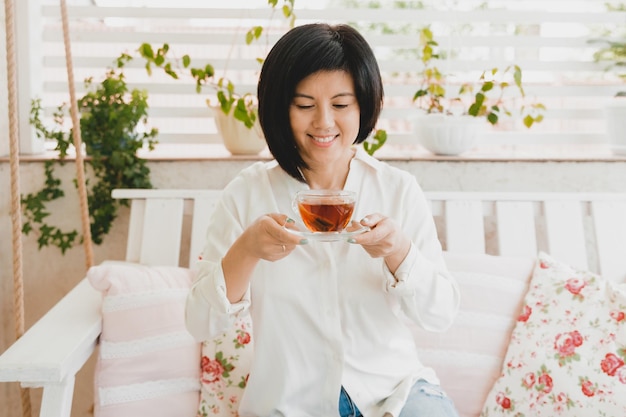 写真 家のテラスでガラスのカップでお茶を飲むアジアの大人の女性のポートレート