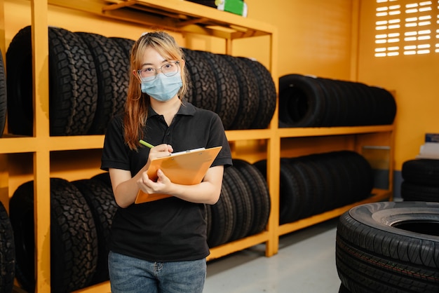 코로나 바이러스나 Covid-19의 확산을 막기 위해 마스크를 쓴 아시아 여성 노동자의 초상화는 창고에서 자동차 타이어 재고를 확인합니다