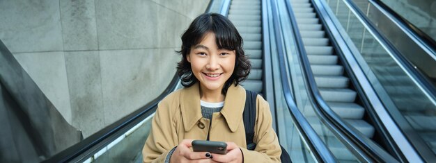 写真 美しいアジアの女子学生の肖像画が街のエスカレーターを下り、携帯電話の使用状況を確認する