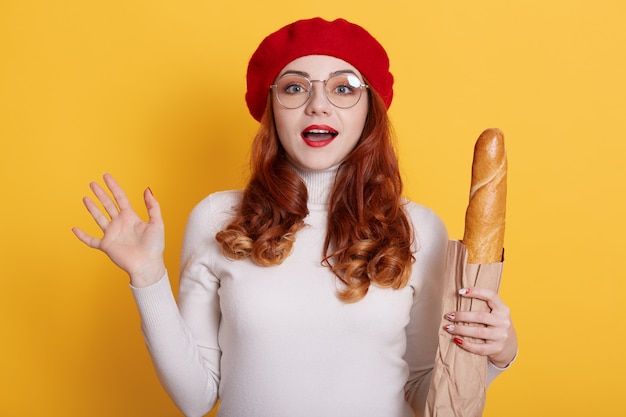 Портрет красивой удивленной молодой женщины, держащей бумажный пакет с хлебным багетом на желтом