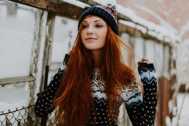 Портрет красивой веснушчатой девушки с длинными рыжими волосами и милой очаровательной улыбкой в серой стильной шляпе, смотрящей и улыбающейся в камеру, стоящую на фоне стены студии изолированный горизонтальный снимок