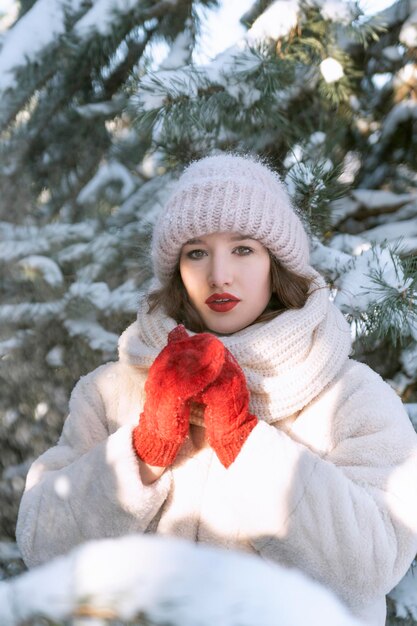 写真 雪に覆われたモミの木の背景に赤いミトンと大きな帽子の美しい少女の肖像画。垂直フレーム。