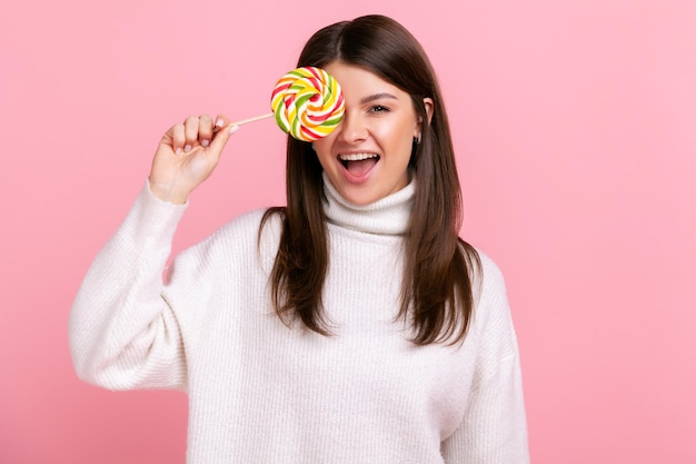 Фото Портрет брюнетки, закрывающей глаз большой вкусной конфетой, смотрящей в камеру с высунутым языком